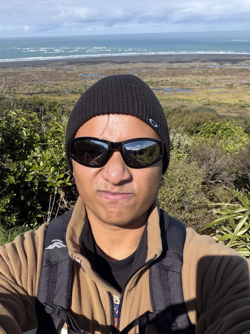 Aaditha hiking, in a beanie and sunglasses with the coast behind