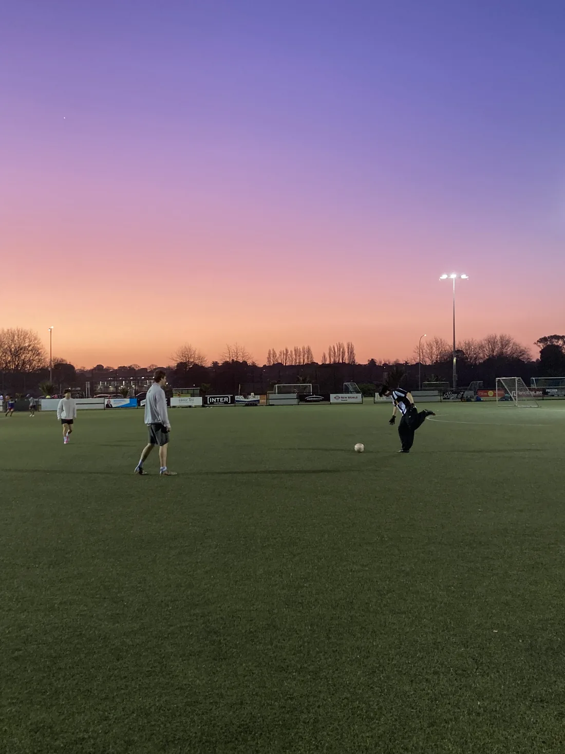 A game of football on a pitch at dusk under a pink sky