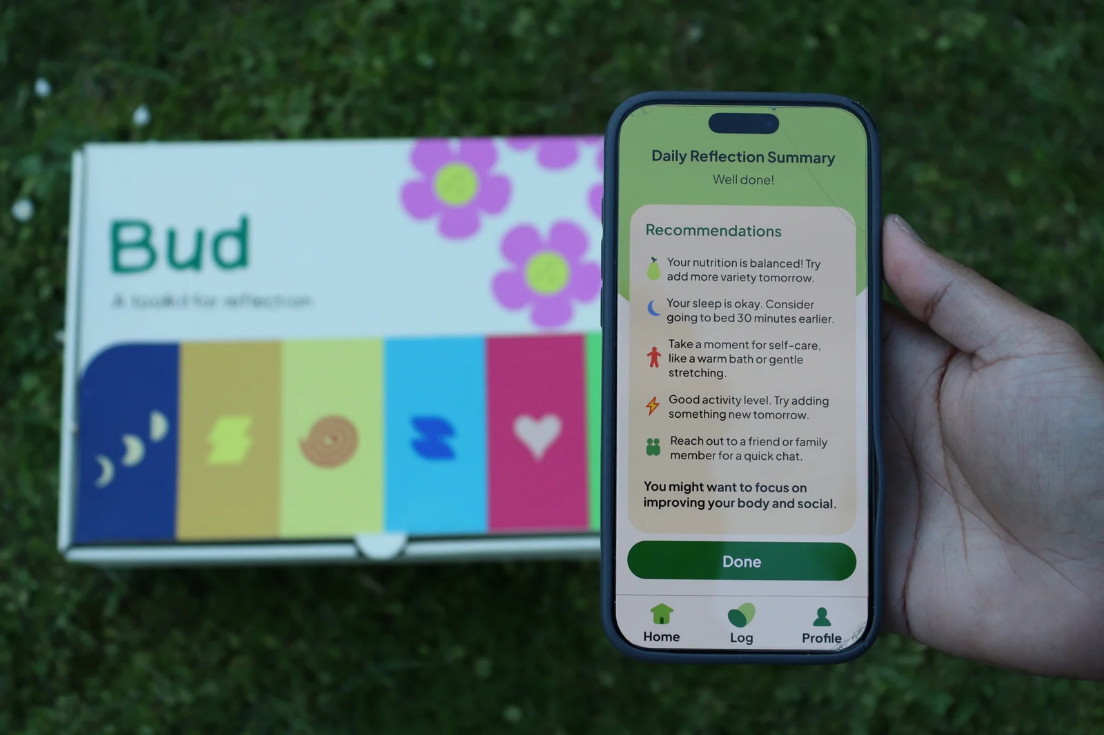 A hand holding a phone showing the Daily Reflection Summary beside the Bud card box on grass